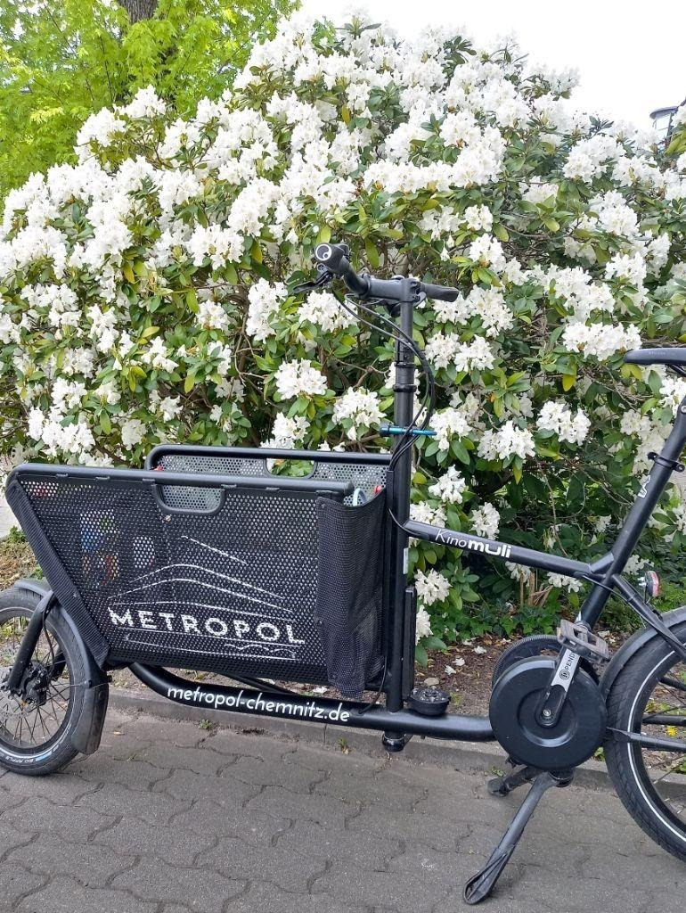 Schwarzes Lastenrad Muli mit Schriftzug "Metropol&quot; vor Fliederbusch